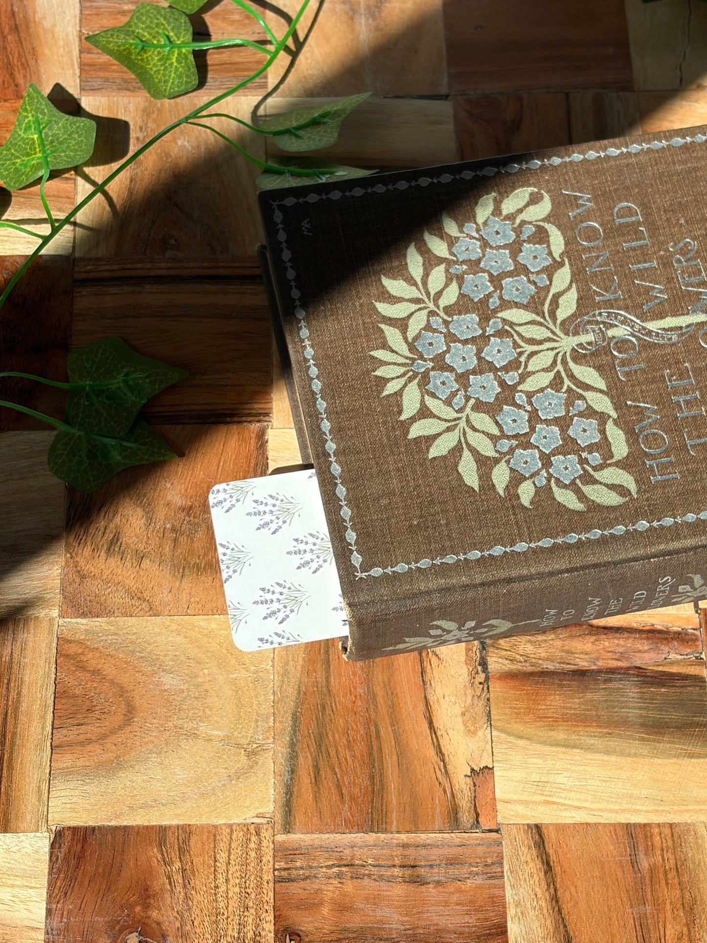 White bookmark with lavender plant bundles in a pattern covering the background, sticking out the top of a book. The book is sitting on top of a wooden background.