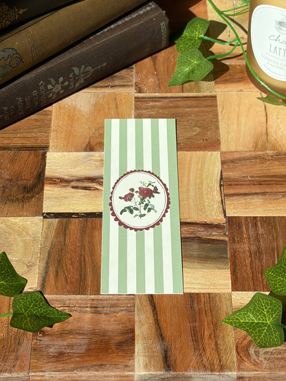 A green striped bookmark with a red round frame picturing red carnations in the center. The bookmark is sitting on a wooden background. 