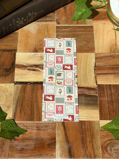 A bookmark with a variety of red, green, and cream stamp designs arranged in a pattern. Featuring a minimal logo at the bottom.The bookmark is sitting on a wooden background.