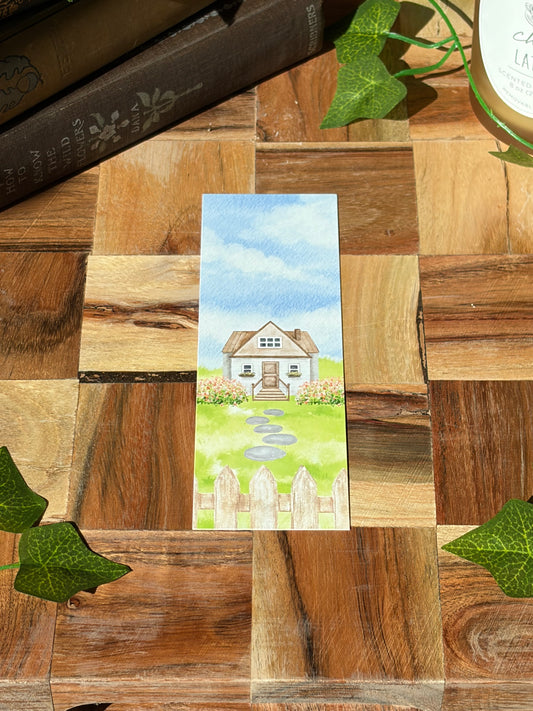 A watercolor landscape design with a picket fence, flowers, stone path, and cottage in the center. The bookmark is sitting on a wooden background.