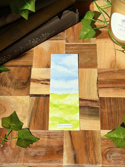 A watercolor landscape design showing sky and grass. Featuring a minimal logo at the bottom. The bookmark is sitting on a wooden background.