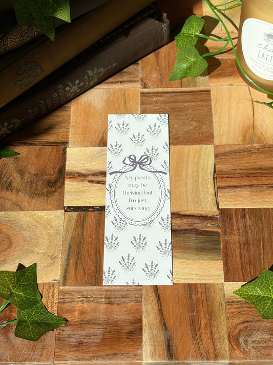 White bookmark with lavender plant bundles in a pattern covering the background. Featuring an oval frame in the center saying "My plants may be thriving but I'm just surviving." The bookmark is sitting on a wooden background. 