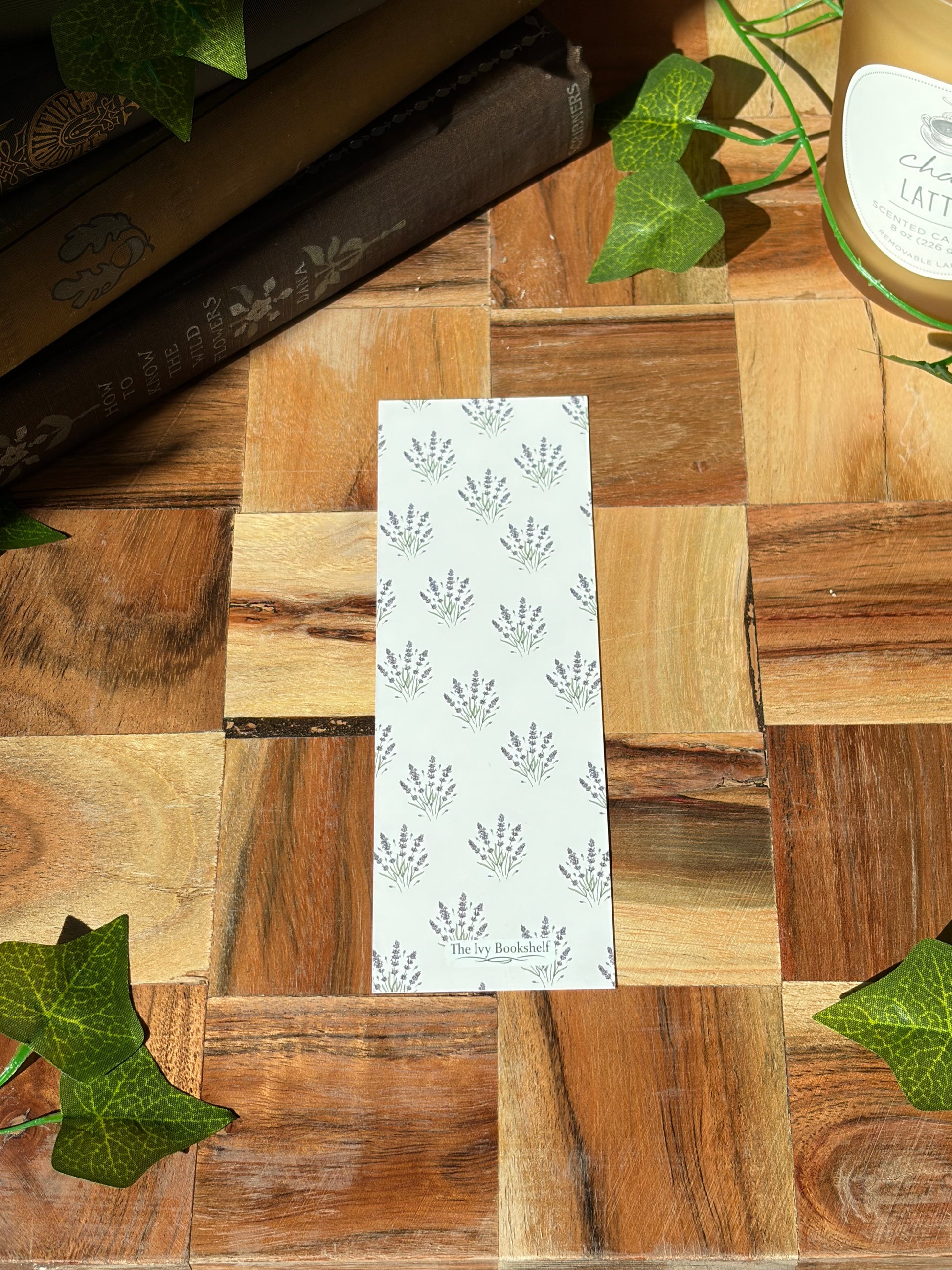 Back of white bookmark with lavender plant bundles in a pattern covering the background. Featuring a minimal logo. The bookmark is sitting on a wooden background. 