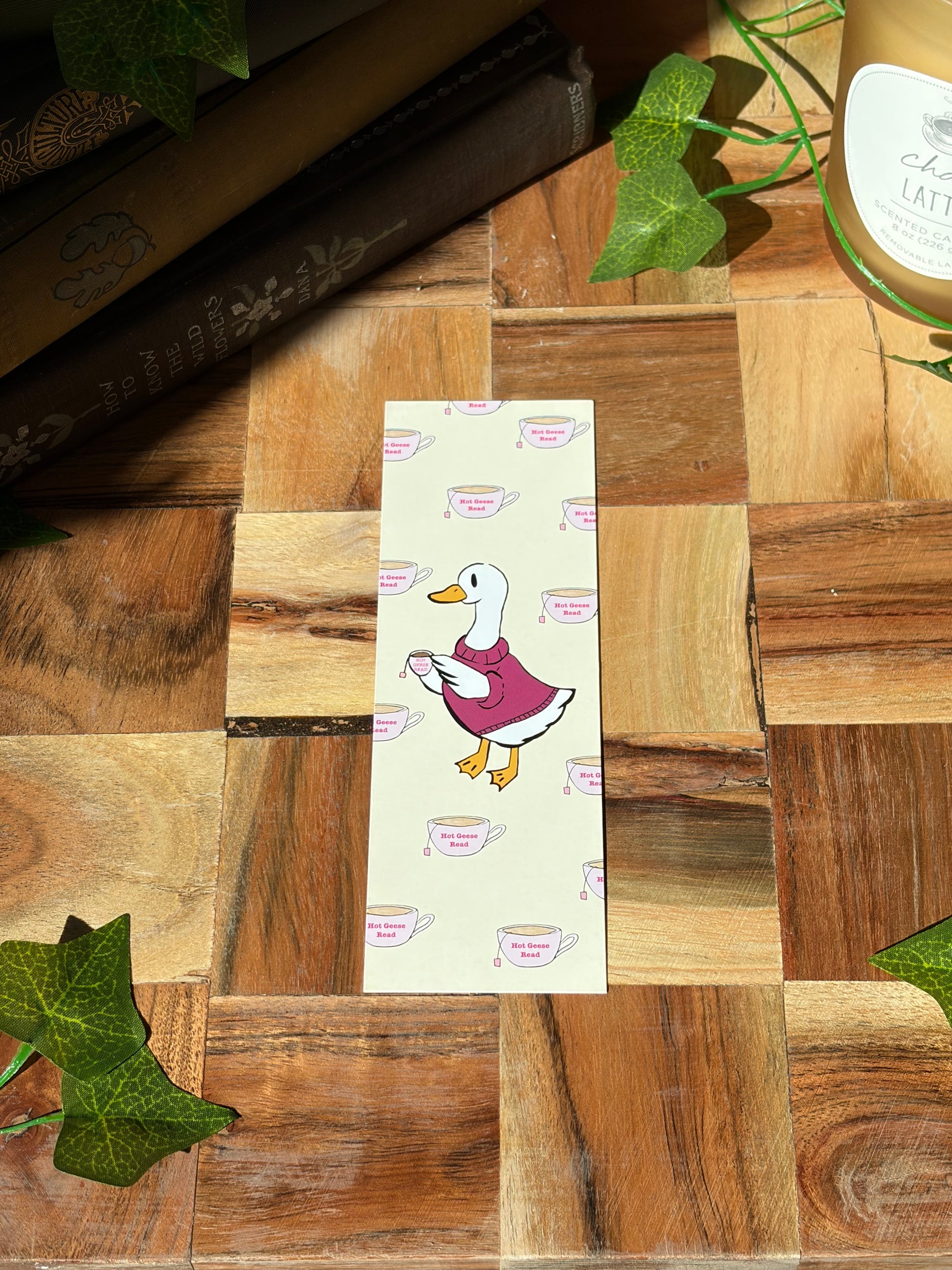Yellow bookmark with a duck in a sweater holding a cup of tea. The background of the bookmark is a pattern of tea cups that say "Hot Geese Read". The bookmark is sitting on a wooden background. 