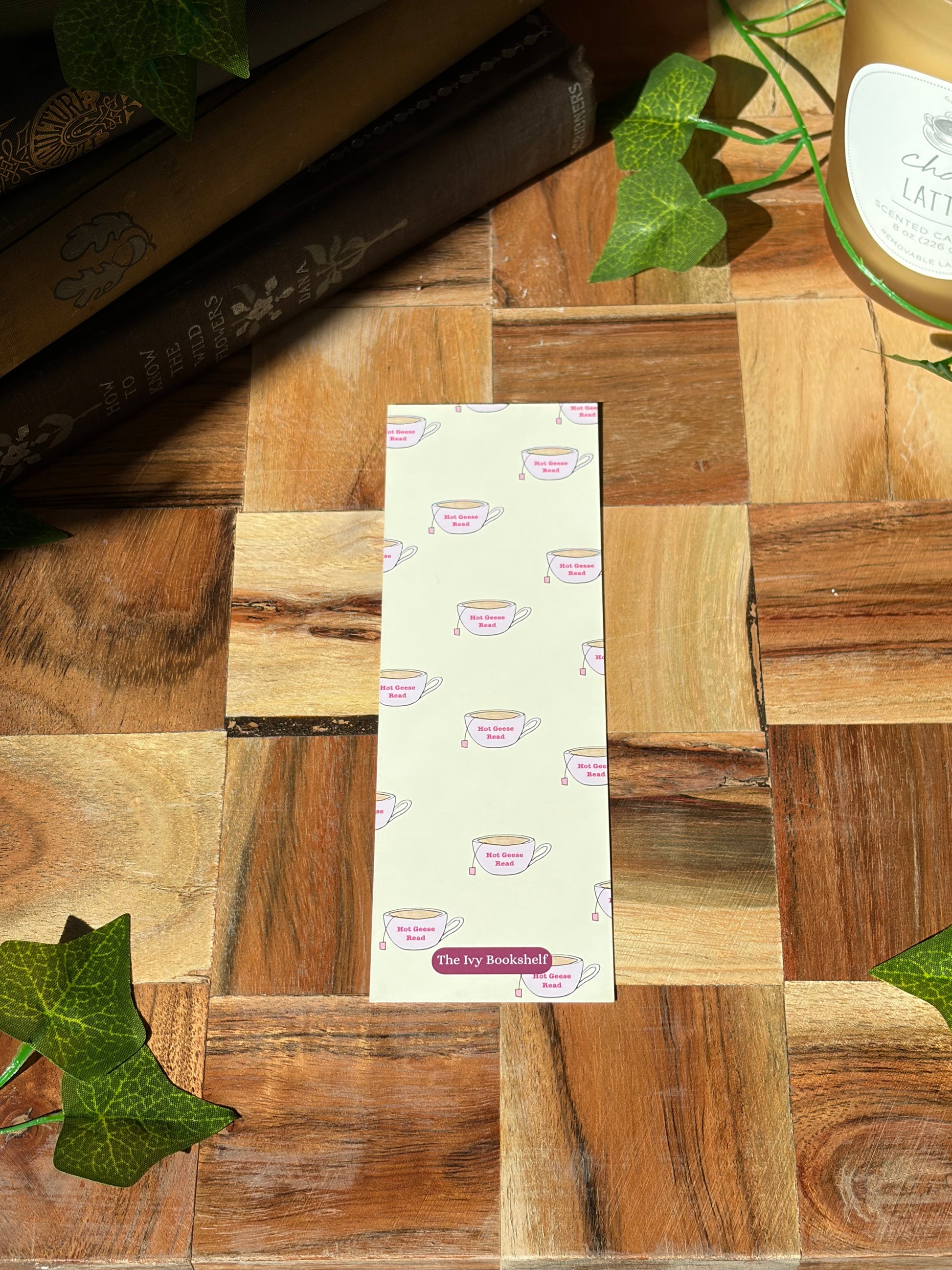 Yellow bookmark featuring a minimal logo at the bottom with a duck in a sweater holding a cup of tea. The background of the bookmark is a pattern of tea cups that say "Hot Geese Read". The bookmark is sitting on a wooden background. 