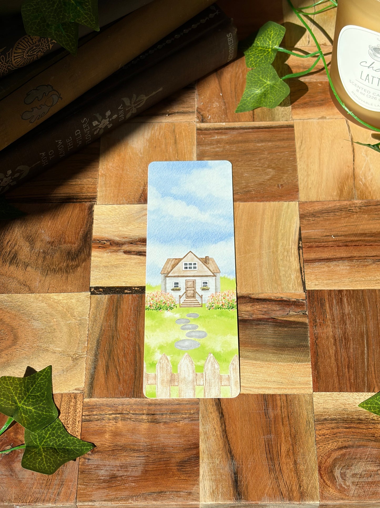 A watercolor landscape design with a picket fence, flowers, stone path, and cottage in the center. Featuring the option for rounded corners. The bookmark is sitting on a wooden background.