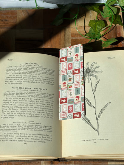 A bookmark with a variety of red, green, and cream stamp designs arranged in a pattern. The bookmark is sitting in an open book. 