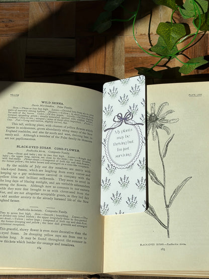 White bookmark with lavender plant bundles in a pattern covering the background. Featuring an oval frame in the center saying "My plants may be thriving but I'm just surviving." The bookmark is sitting on an open book.