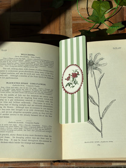 A green striped bookmark with a red round frame picturing red carnations in the center. The bookmark is sitting in an open book.