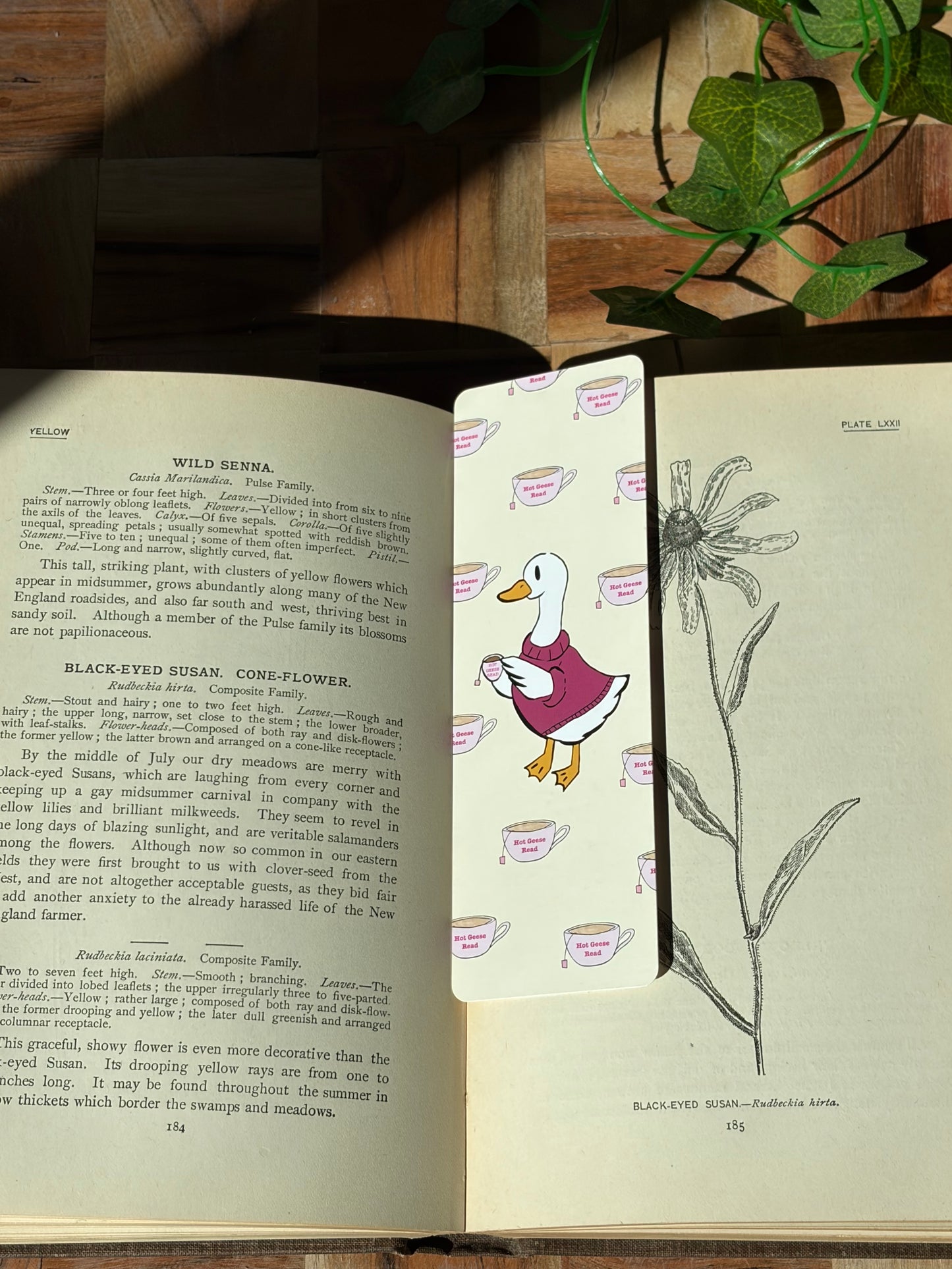 Yellow bookmark with a duck in a sweater holding a cup of tea. The background of the bookmark is a pattern of tea cups that say "Hot Geese Read". The bookmark is sitting in an open book. 