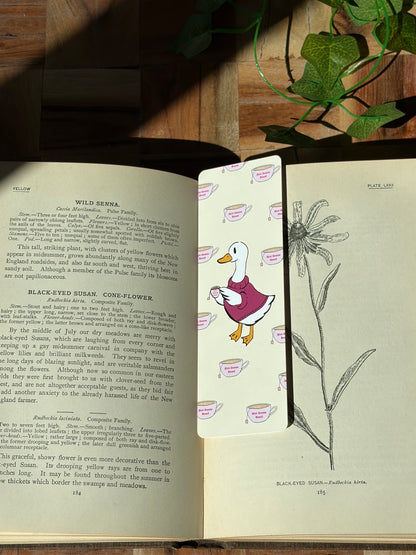Yellow bookmark with a duck in a sweater holding a cup of tea. The background of the bookmark is a pattern of tea cups that say "Hot Geese Read". The bookmark is sitting in an open book. 