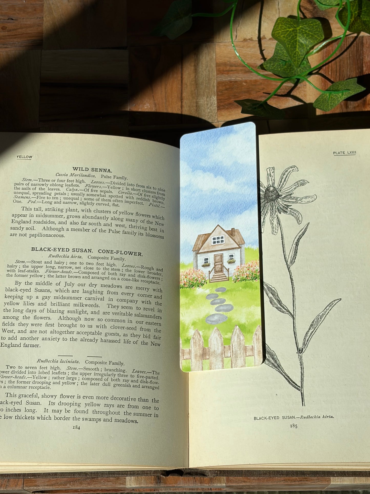 A watercolor landscape design with a picket fence, flowers, stone path, and cottage in the center. The bookmark is sitting inside an open book.
