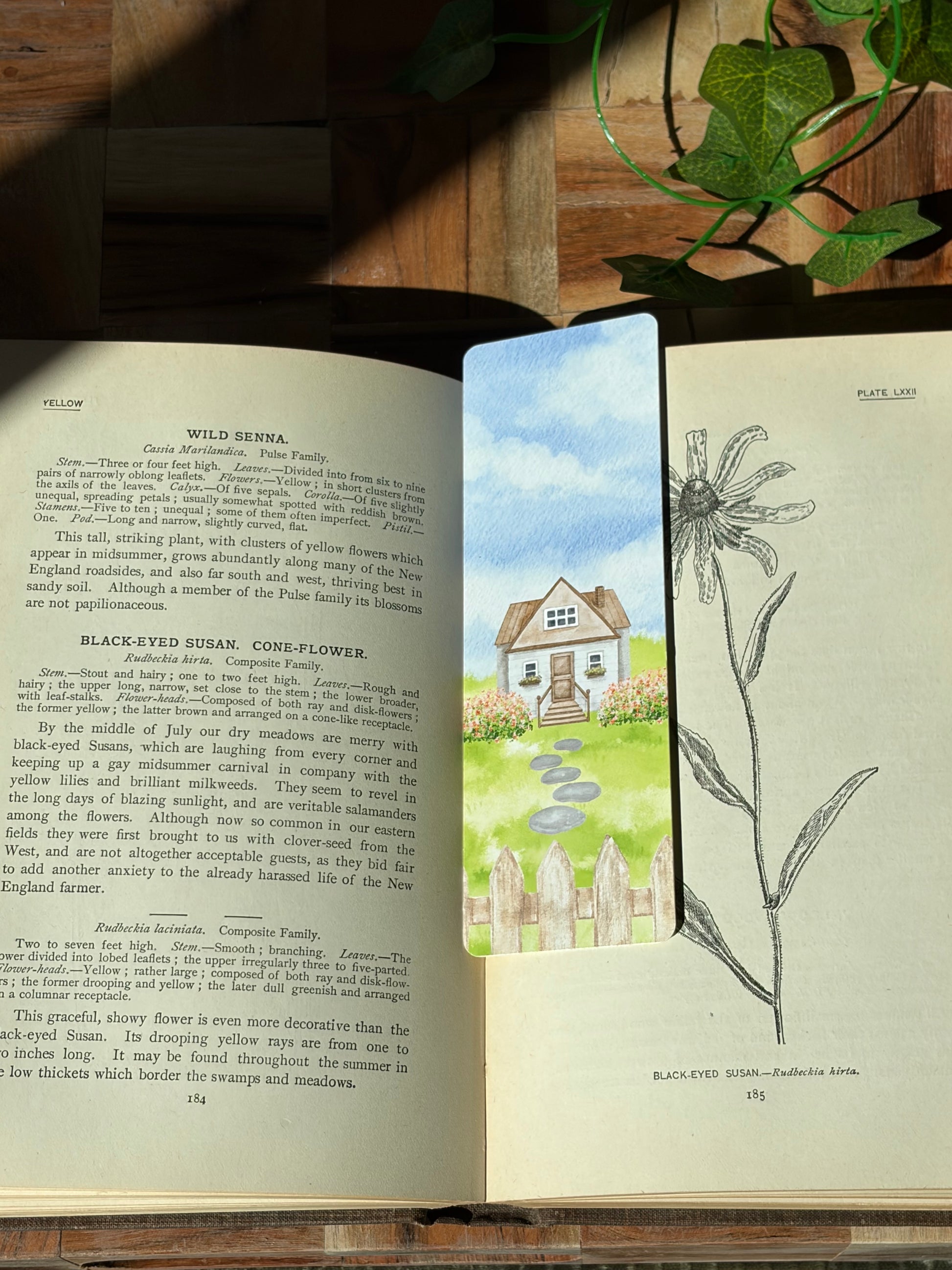 A watercolor landscape design with a picket fence, flowers, stone path, and cottage in the center. The bookmark is sitting inside an open book.