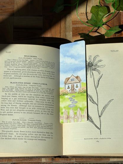 A watercolor landscape design with a picket fence, flowers, stone path, and cottage in the center. The bookmark is sitting inside an open book.