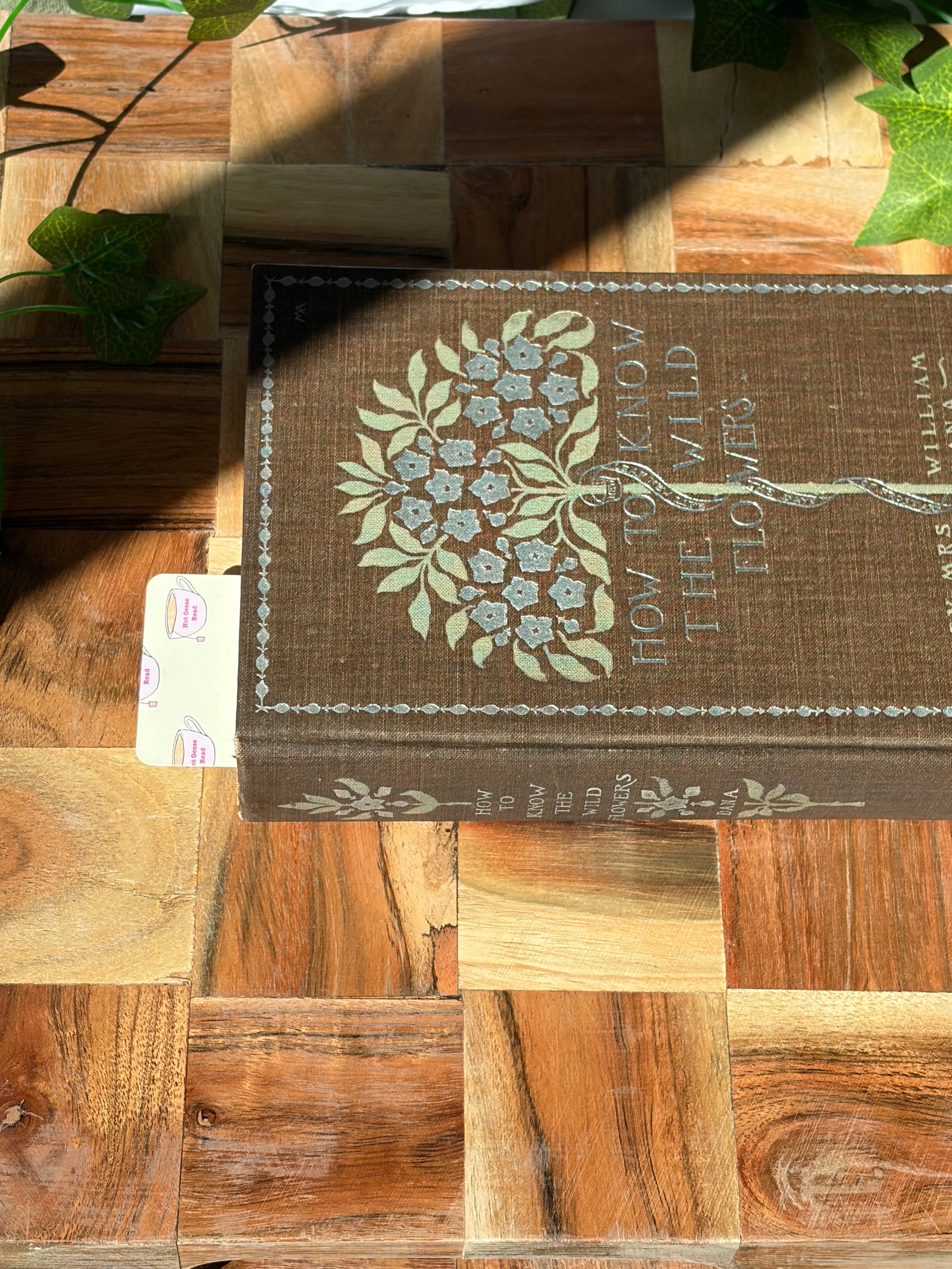 Yellow bookmark with a pattern of tea cups that say "Hot Geese Read". The bookmark is sitting inside a closed book on a wooden background.