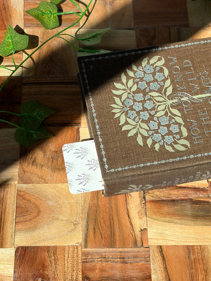 White bookmark with lavender plant bundles in a pattern covering the background, sticking out the top of a book. The book is sitting on top of a wooden background.