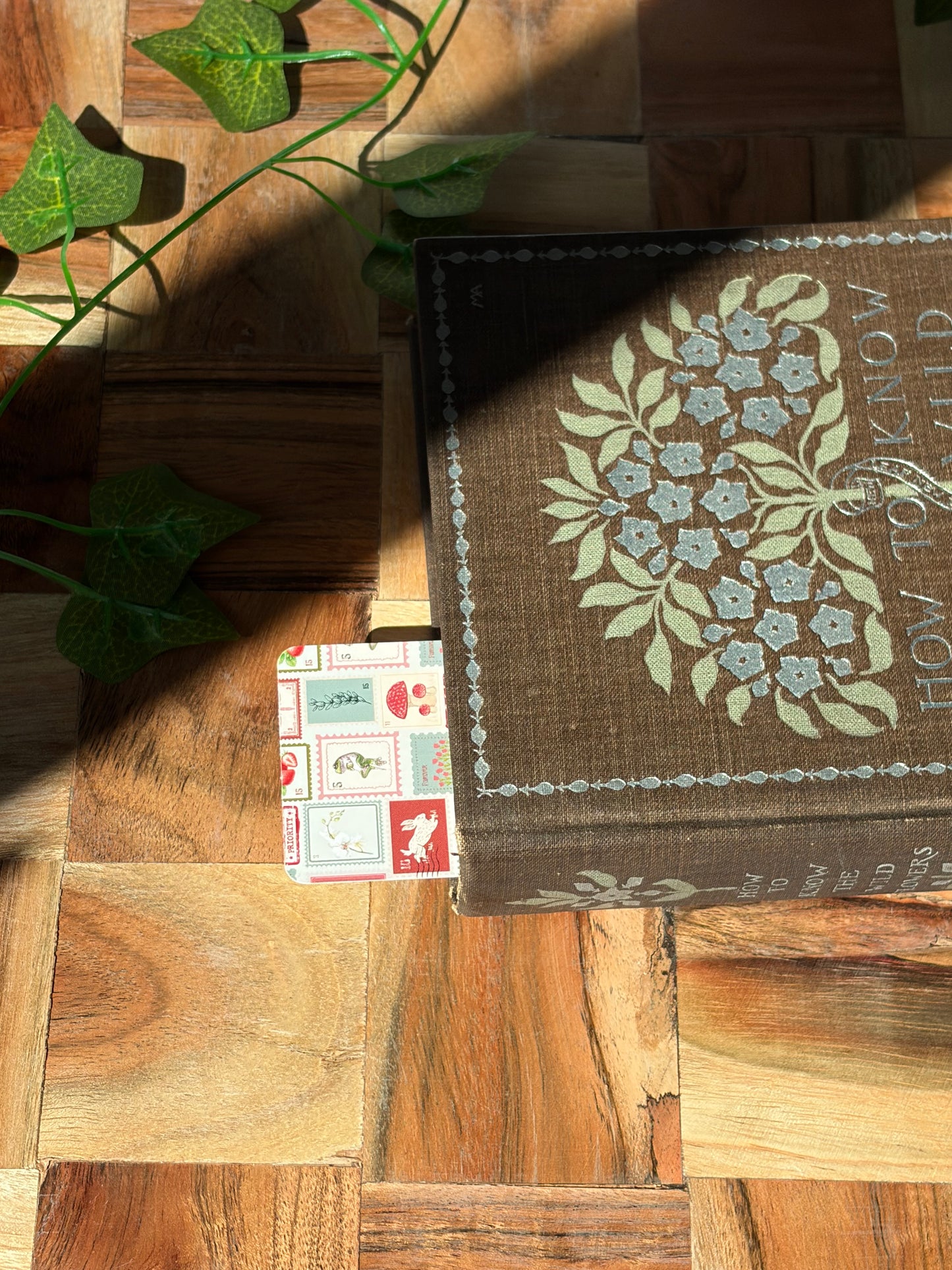 A bookmark with a variety of red, green, and cream stamp designs arranged in a pattern. The bookmark is sticking out the top of a closed book on a wooden background.